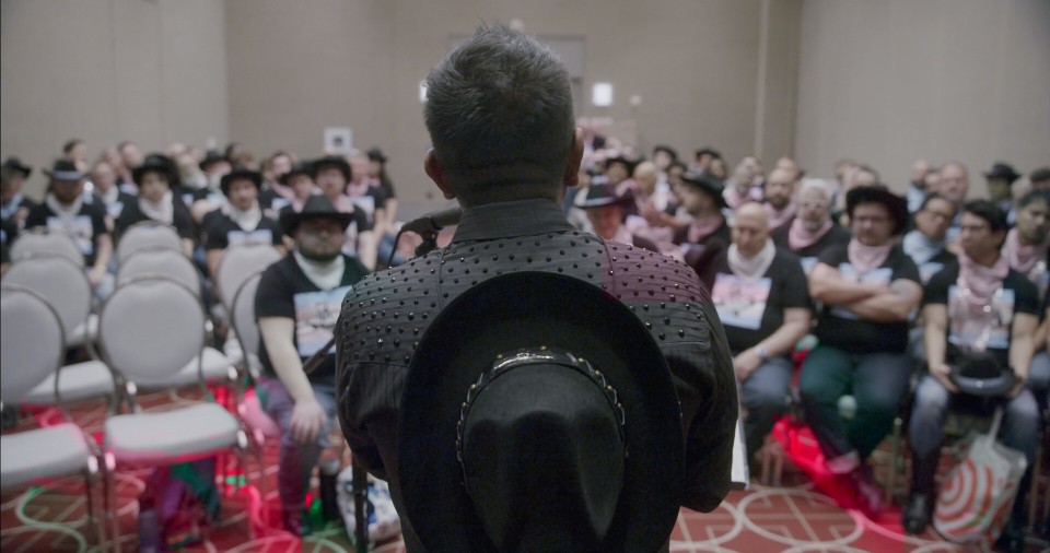 A performer in a studded black shirt holding a cowboy hat, facing the Austin Gay Men's Chorus
