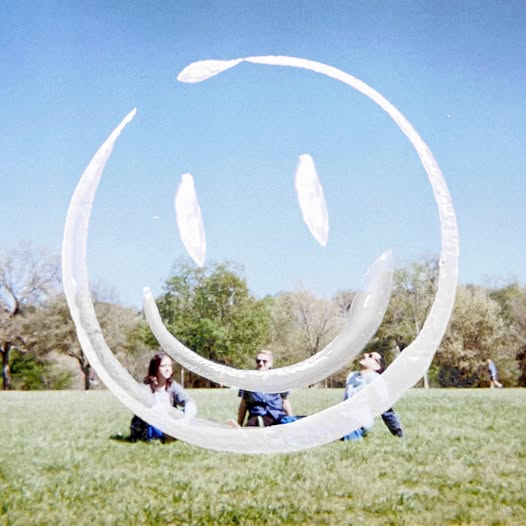 Painterly album cover: a white smiley face drawn over a photo of the band in a park