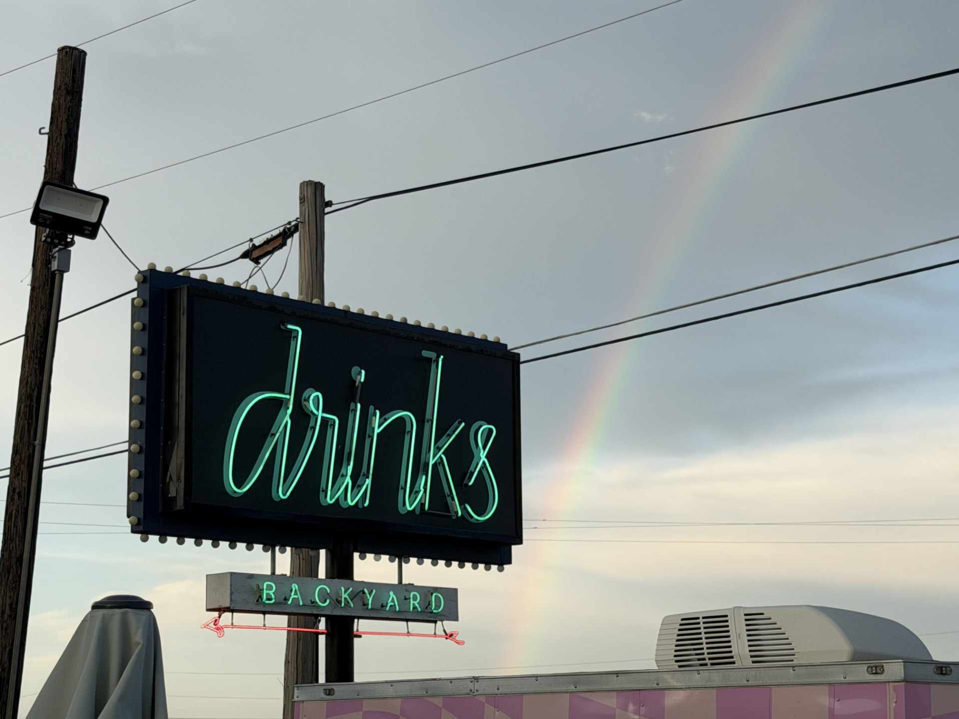 Neon Drinks Backyard sign with a rainbow in the sky behind it
