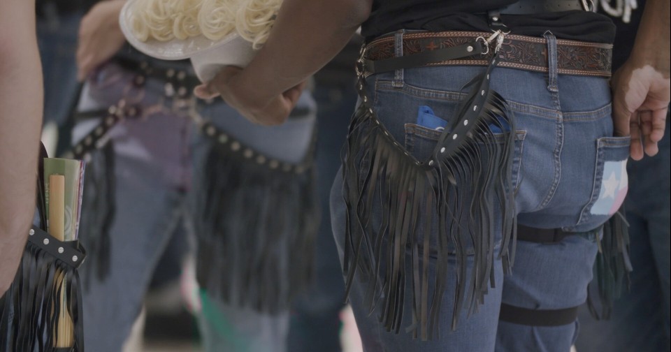 Close-up of chorus members' black fringe sashes over jeans, one with a trans flag patch