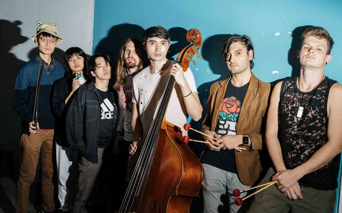 The eight members of Austin band Maru Haru with strings, upright bass and guitar against a teal wall