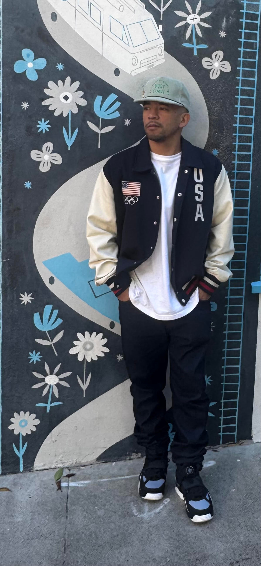 Bboy Wicket in a USA varsity jacket standing in front of a floral Austin mural