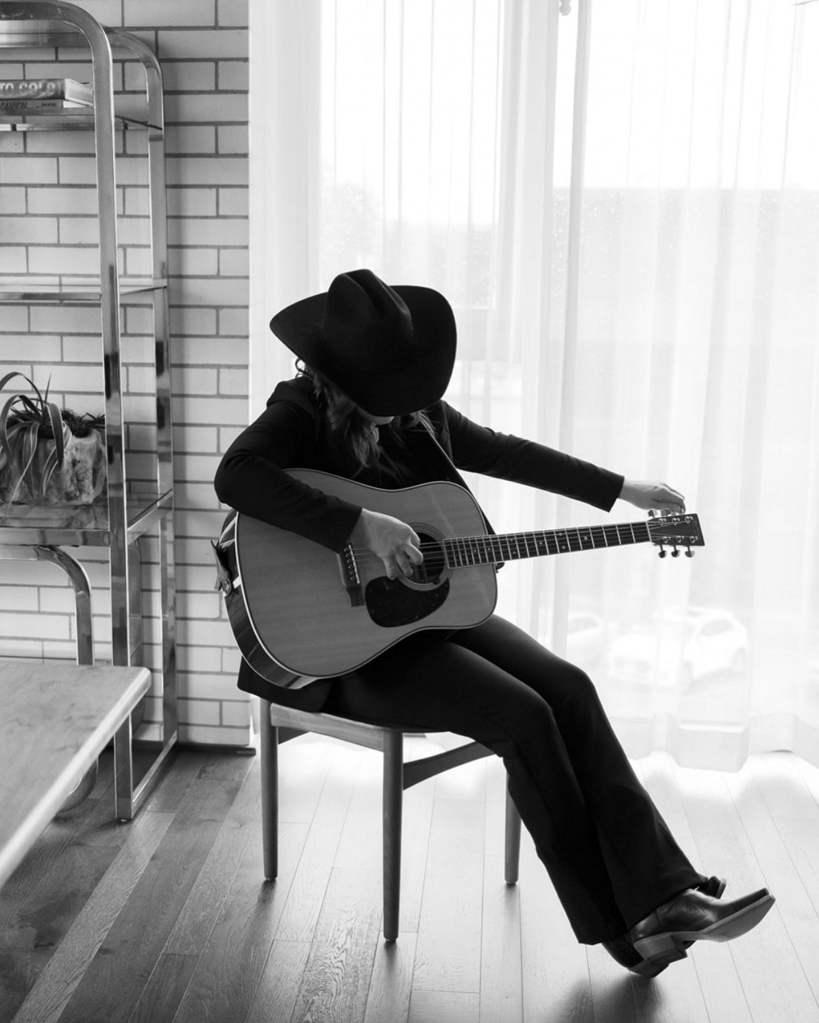 Black and white photo of Steph Cash seated playing acoustic guitar by a window