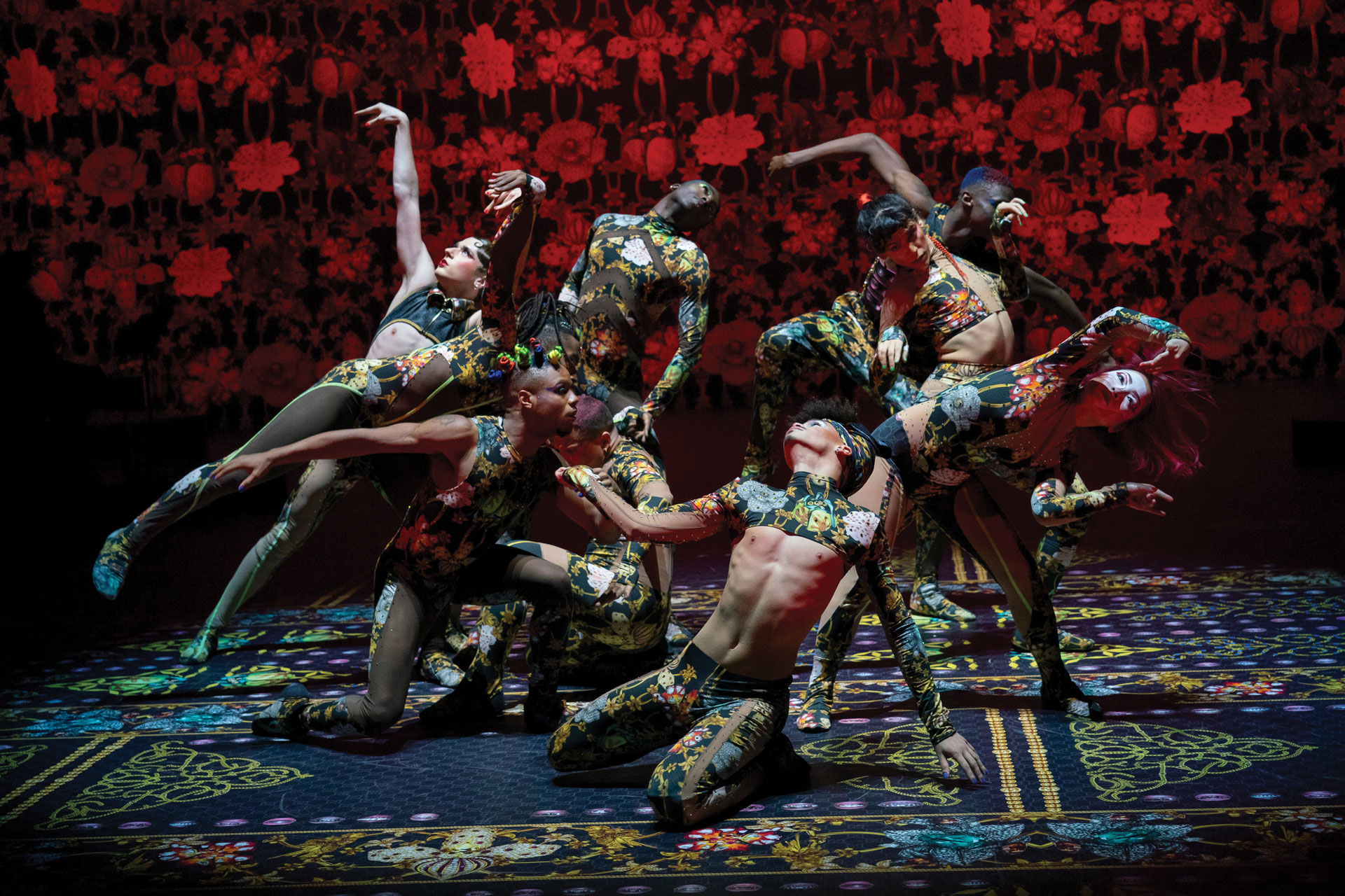 Dancers in ornate patterned costumes posed mid-movement against a red floral projection (credit: Stephanie Berger)