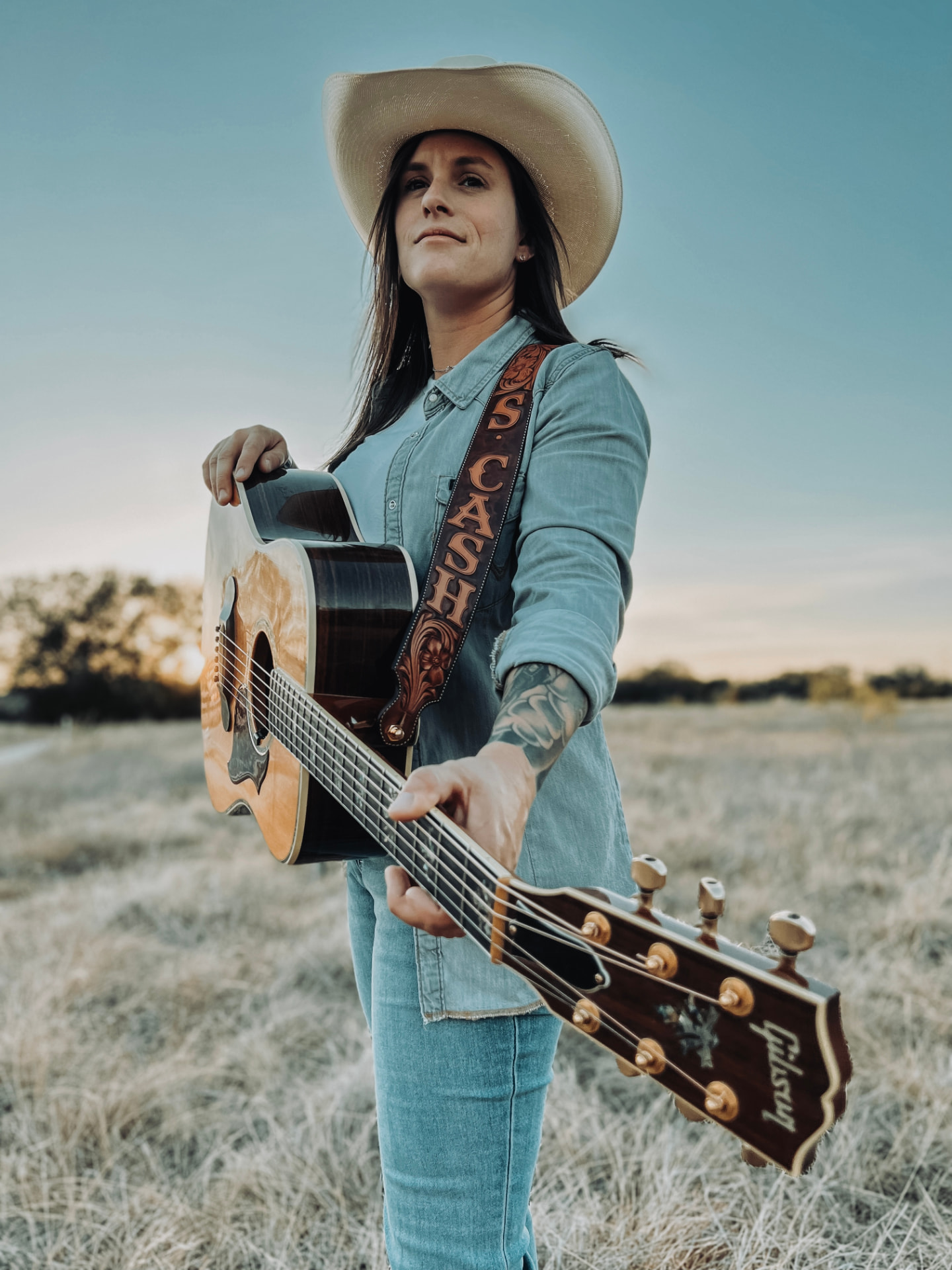 Steph Cash in a straw cowboy hat holding a Gibson acoustic guitar in a field at sunset